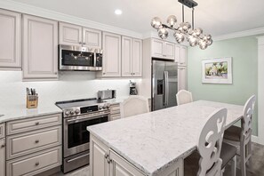 Una cocina moderna con armarios blancos, electrodomésticos de acero inoxidable, encimera de mármol y un comedor con sillas blancas.