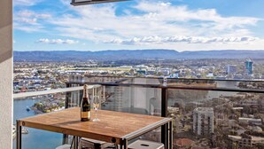 A balcony with a wooden table, a bottle of wine, and a glass. The view includes a cityscape with mountains in the distance. - Circle on Cavill River View (Surfers Paradise)