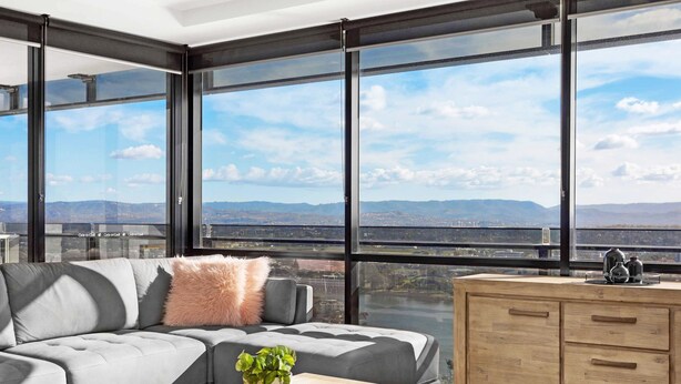 A modern living room with a grey sofa, a wooden coffee table, and a view of a cityscape through large windows. - Circle on Cavill River View (Surfers Paradise)