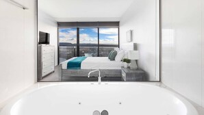 A modern bathroom with a large bathtub, a view of a bedroom with a bed and a window, and a white cabinet with a television. - Circle on Cavill River View (Surfers Paradise)