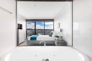 A modern bathroom with a large bathtub, a view of a bedroom with a bed and a window, and a white cabinet with a television. - Circle on Cavill River View (Surfers Paradise)