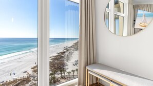 A room with a large window overlooking a beach, a round mirror on the wall, and a bench. - Gulf Front Balcony, Gorgeous Views, Near Destin Harbor (Destin)