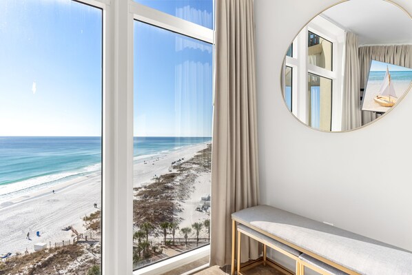 A room with a large window overlooking a beach, a round mirror on the wall, and a bench.
