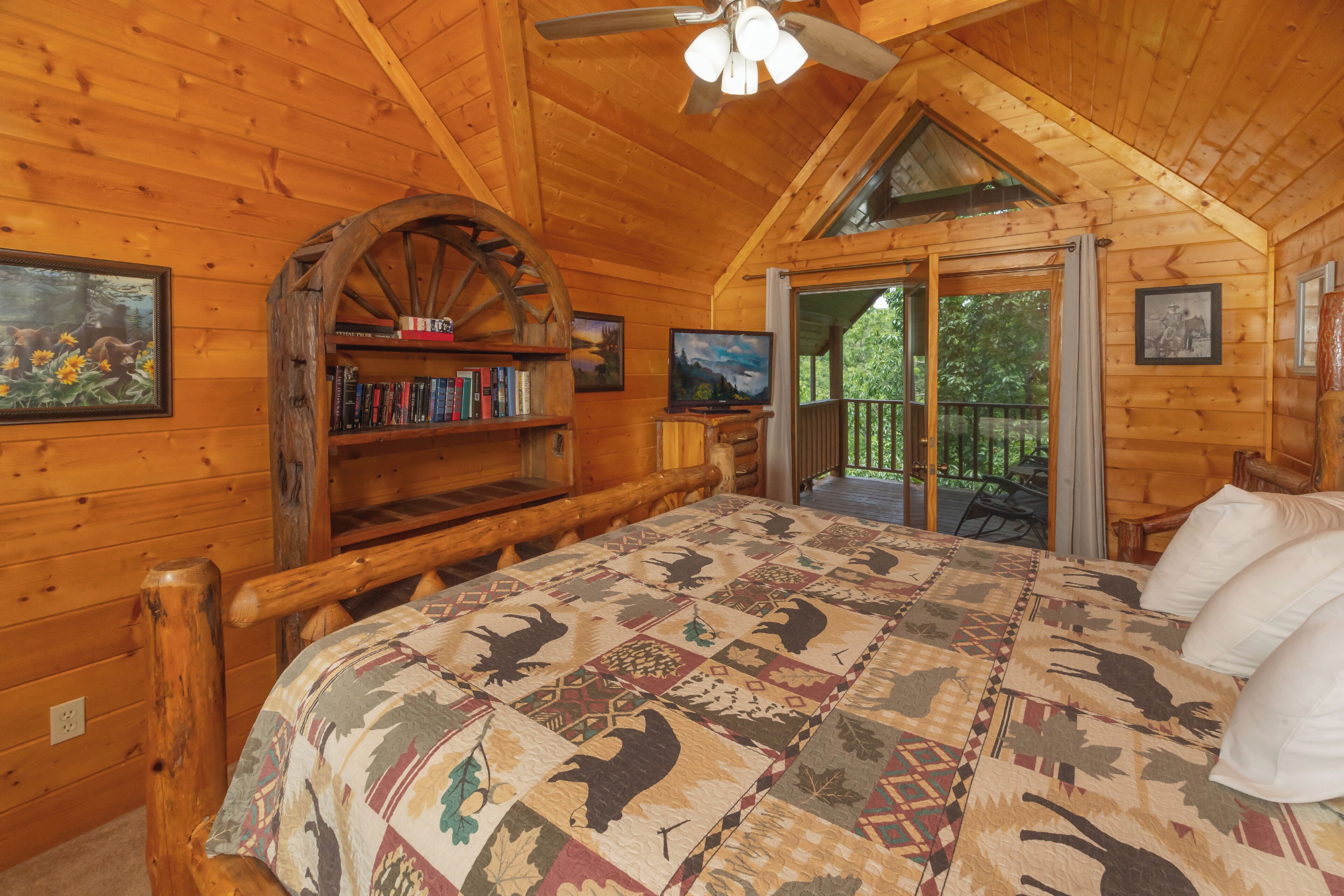 Een slaapkamer in een blokhut met een groot bed, houten boekenplanken, een plafondventilator en een balkon met uitzicht op de omgeving.
