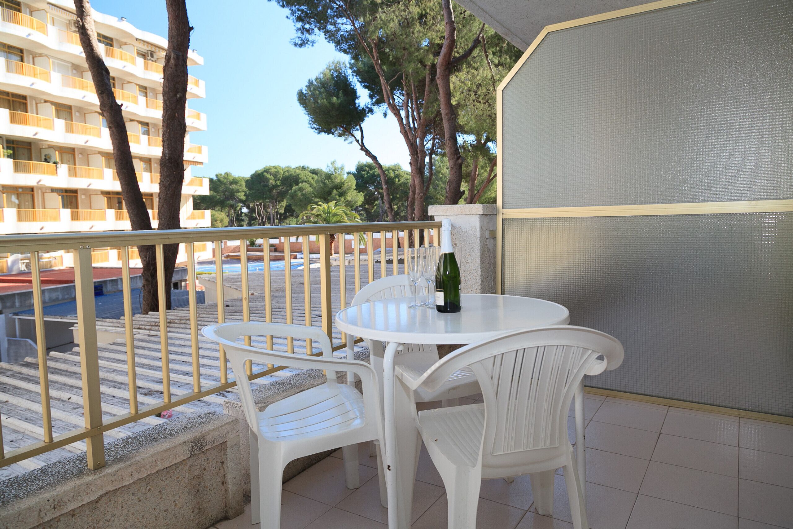 Een balkon met een witte tafel en stoelen, een fles wijn en uitzicht op een gebouw en bomen.