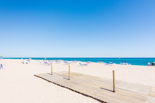 A beach with a wooden pier, blue lounge chairs, and a clear blue sky.