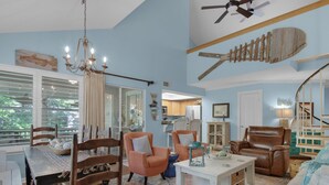 A spacious living room with a large window, a chandelier, a ceiling fan, and a wooden fish decoration. - Bright and Cheerful Cottage, Updated Kitchen, Large Flat Screen TV (Miramar Beach)