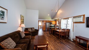 A spacious living room with a brown sofa and wooden dining table, a kitchen with wooden cabinets, and a large window providing natural light.