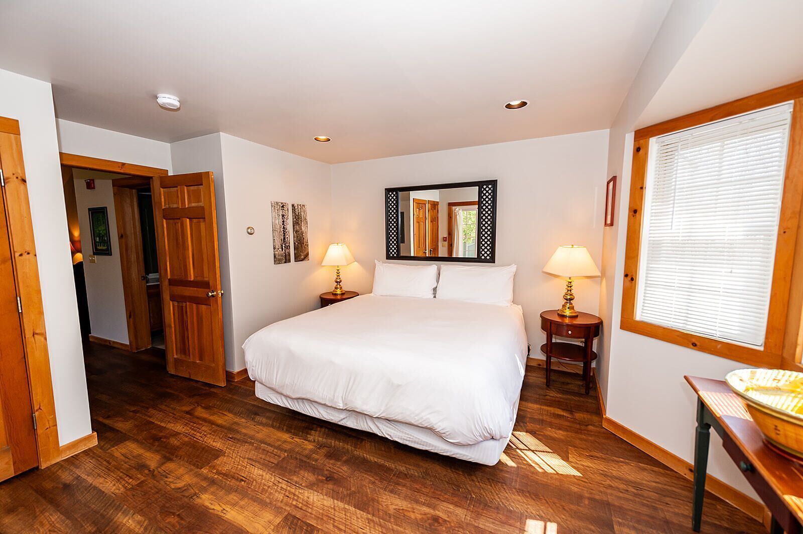 A bedroom with a large bed, two bedside tables with lamps, a wooden floor, a door leading to another room, and a window with blinds.