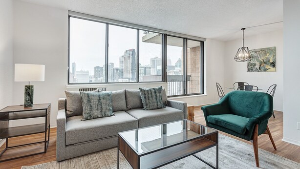 Una sala moderna con un sofá gris, un sillón verde, una mesa de centro de vidrio y vistas a la ciudad a través de amplios ventanales.