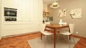 Eine moderne Küche mit weißen Schränken, einem eingebauten Ofen und einem Weinkühlschrank. Ein Esstisch mit vier Stühlen und einem Teppich sind ebenfalls vorhanden.