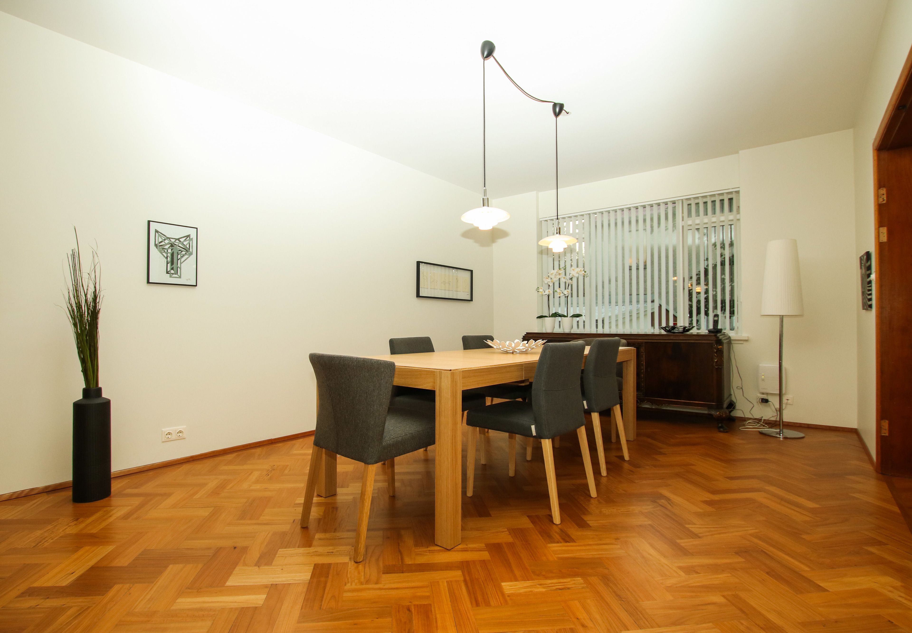 A dining area with a wooden table, six chairs, two pendant lights, and a potted plant.