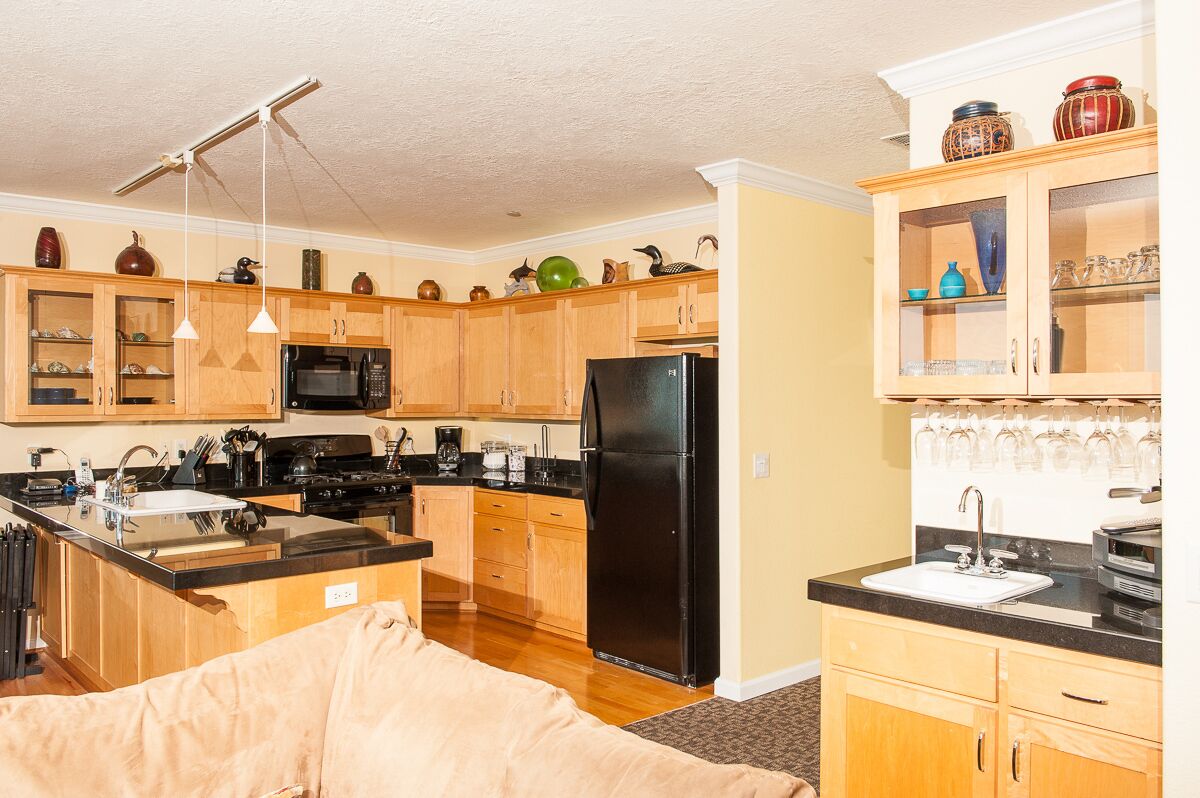 A compact kitchen with wooden cabinets, a black refrigerator, and a microwave. There are hanging lights and a variety of decorative items on the shelves.