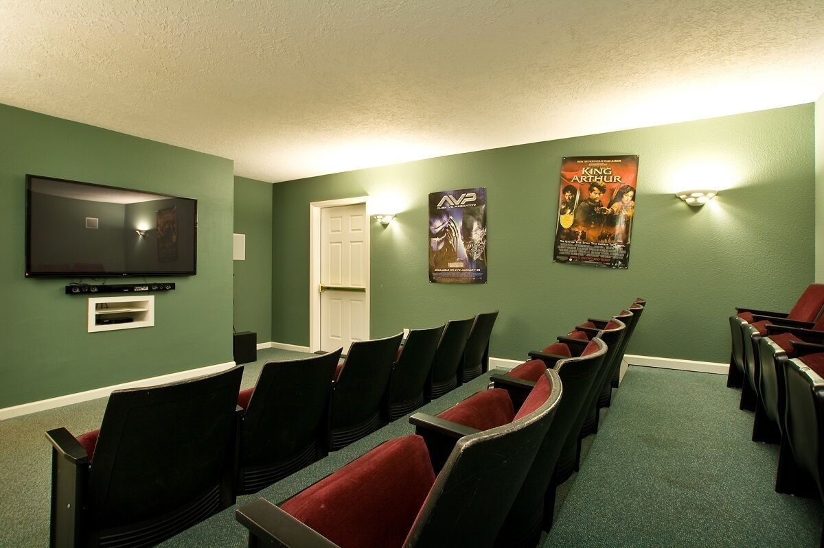 A room with green walls, a flat-screen TV mounted on the wall, and a poster of 'King Arthur'. There are rows of black chairs with red cushions.