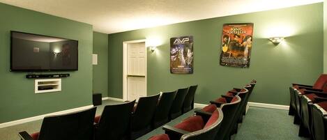 A room with green walls, a flat-screen TV mounted on the wall, and a poster of 'King Arthur'. There are rows of black chairs with red cushions.