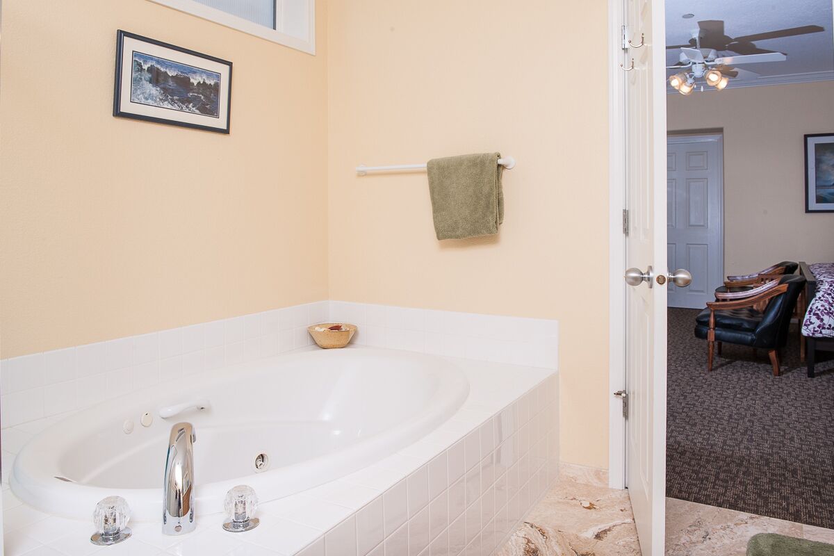A bathroom with a large white bathtub, a white towel rack with a green towel, a framed picture on the wall, and a ceiling fan.