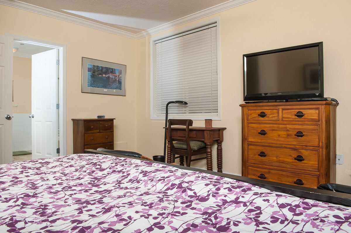 Une chambre à coucher comprenant un lit, une commode, un téléviseur, une chaise et une fenêtre avec des stores.