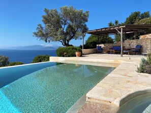 Uma piscina infinita transparente com um deck de pedra, área de estar ao ar livre e vista para o mar e montanhas.
