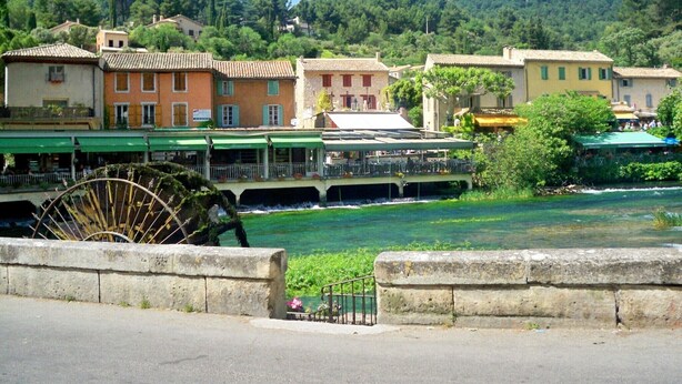 Un pueblo a orillas del río con edificios, una noria grande y un puente.
