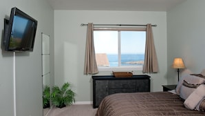 A bedroom with a bed, a television mounted on the wall, a window with curtains, a dresser, and a lamp.