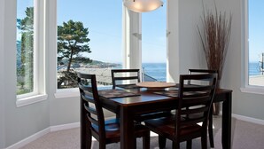 A dining area with a table and chairs, large windows with a view of the ocean and trees, and a hanging light fixture.