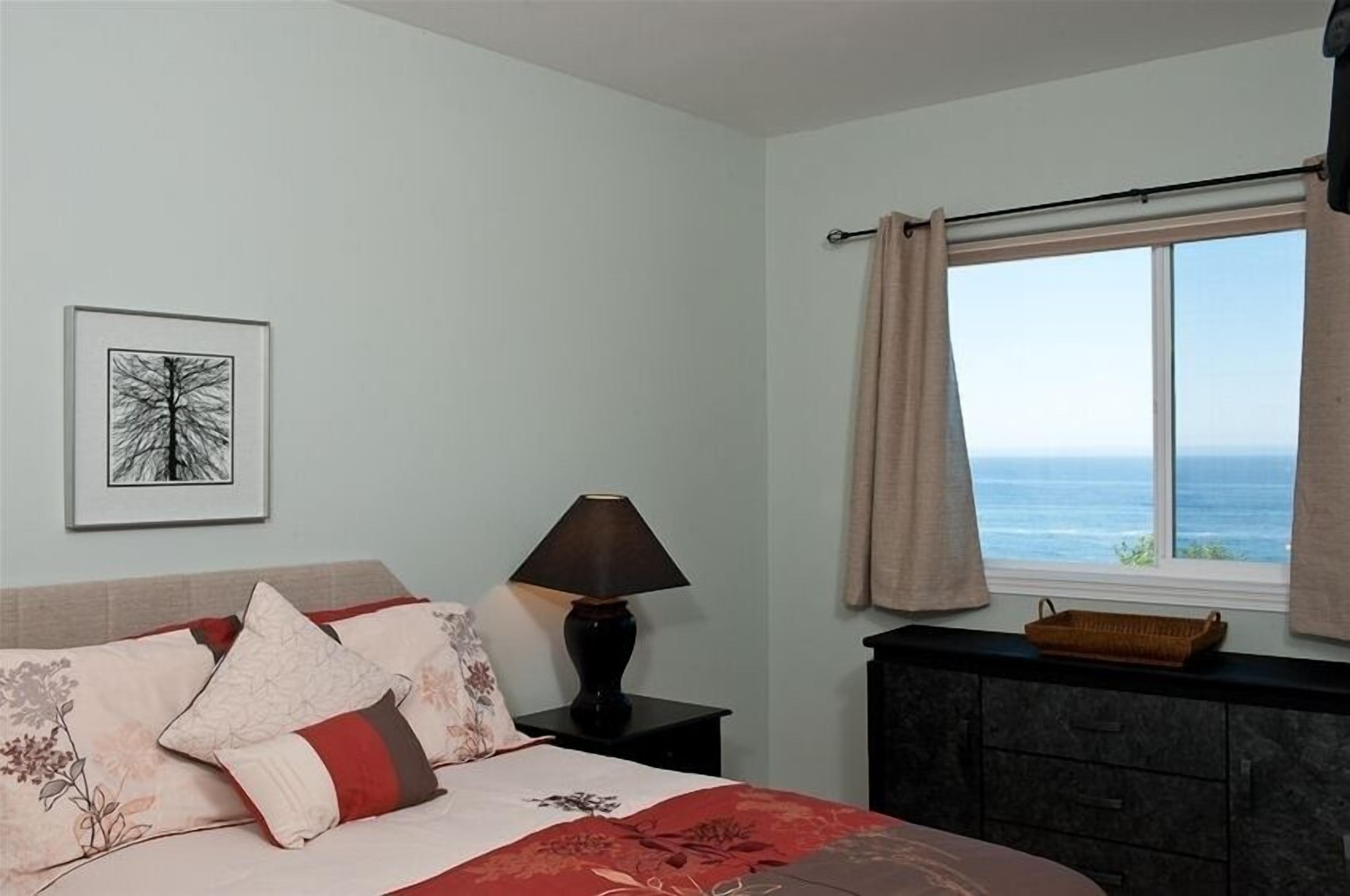 Una camera da letto con un letto, un comodino, una lampada, un quadro incorniciato, una finestra con tende e vista sul mare.