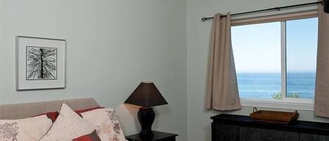 Una camera da letto con un letto, un comodino, una lampada, un quadro incorniciato, una finestra con tende e vista sul mare.