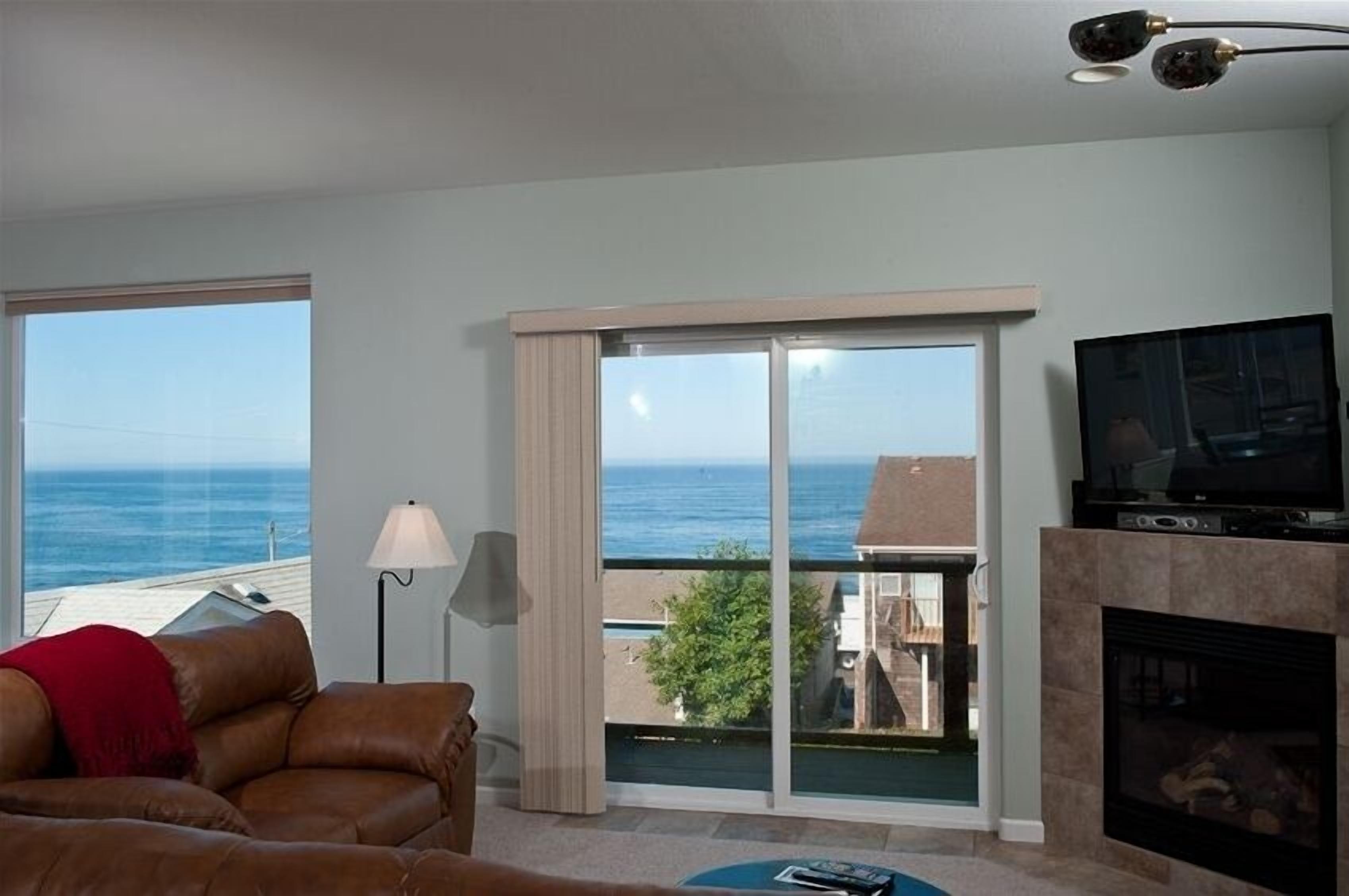 A living room with a large window overlooking the sea, a fireplace, and a television.