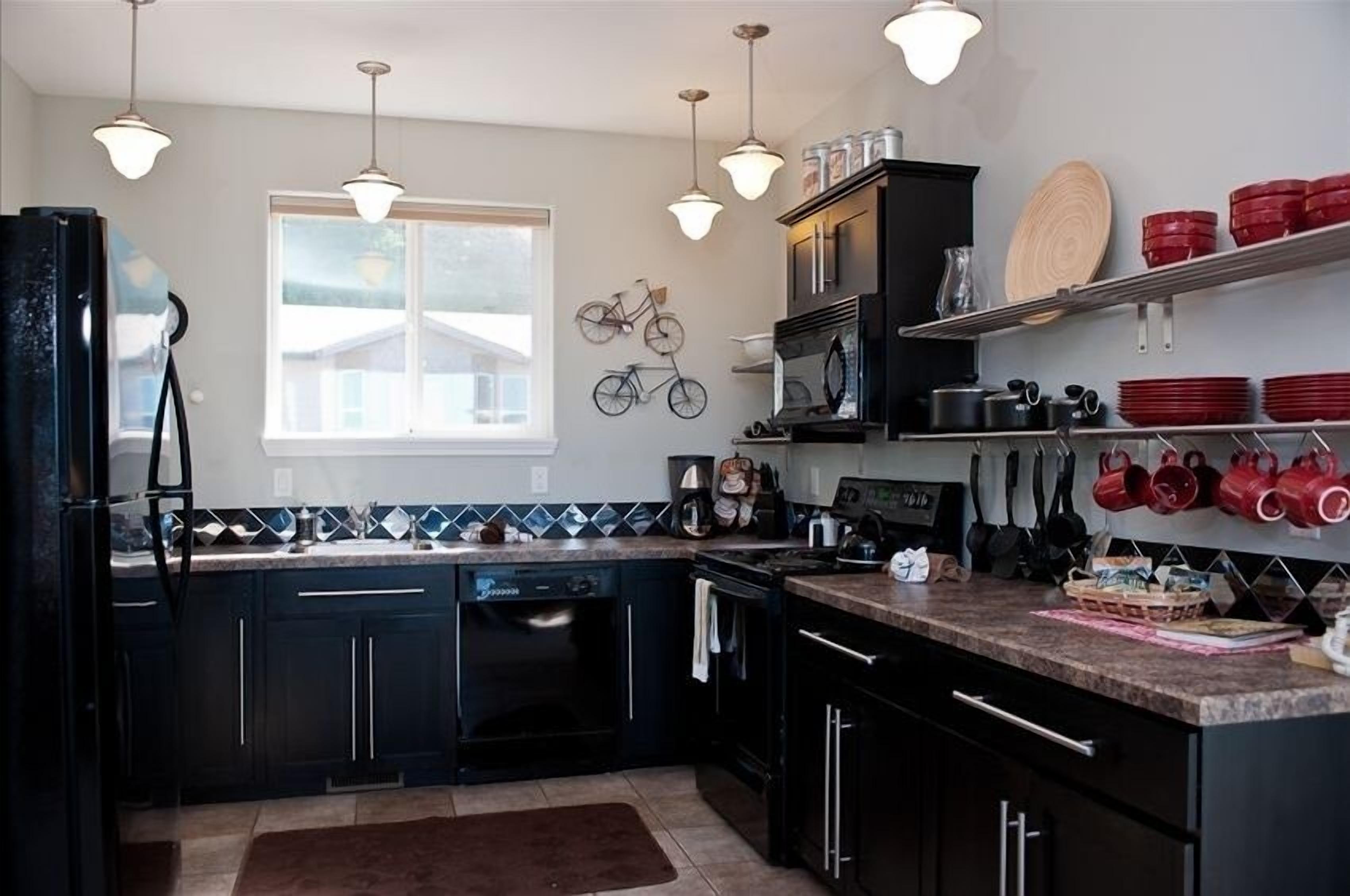 A modern kitchen with black cabinets, a dishwasher, a stove, and a microwave. There are hanging pots and pans, and a window providing natural light.