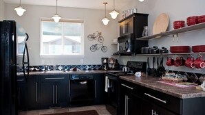 A modern kitchen with black cabinets, a dishwasher, a stove, and a microwave. There are hanging pots and pans, and a window providing natural light.