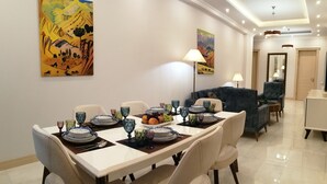 Ein Essbereich mit einem für vier Personen gedeckten Tisch, umgeben von Stühlen. An den Wänden befinden sich zwei Gemälde, ein Sofa und im Hintergrund ein Fernseher.