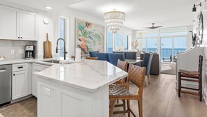 A modern kitchen with white cabinetry, a marble island, and a dining area with wicker chairs. - Gulf Front, Corner Condo, Sunrise Views, Pool (Destin)