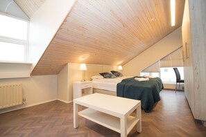 Um quarto com teto inclinado, uma cama com almofadas, uma mesa de cabeceira com abajur, uma cadeira e uma janela.