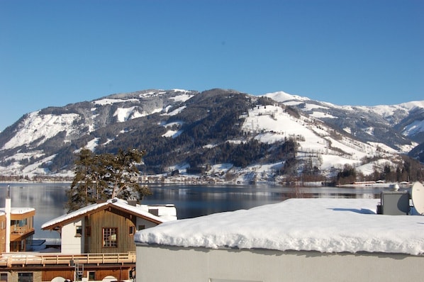 A chalet-style building with a wooden deck and a snow-covered roof, overlooking a lake and mountains.