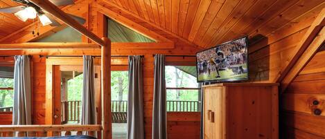 A wooden cabin room with a bed, a television on a wooden cabinet, and curtains on the windows.