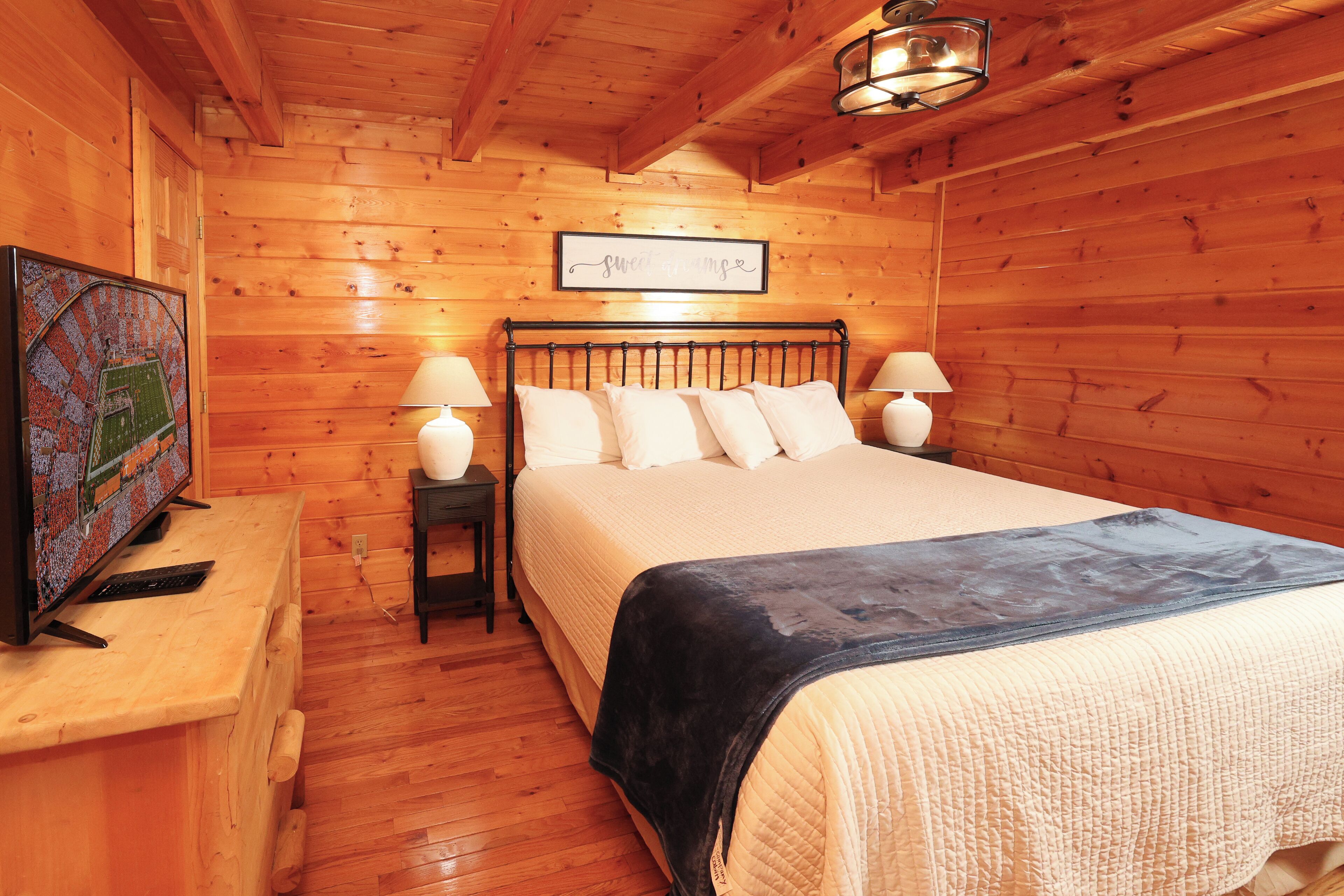 Ein Schlafzimmer einer Holzhütte mit einem Bett, zwei Nachttischlampen, einem Fernseher auf einer Holzkommode und einem gerahmten Bild an der Wand.