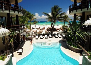 Un espace piscine aménagé dans un complexe hôtelier, avec des chaises longues et des palmiers, offrant une vue sur une plage et l’océan.
