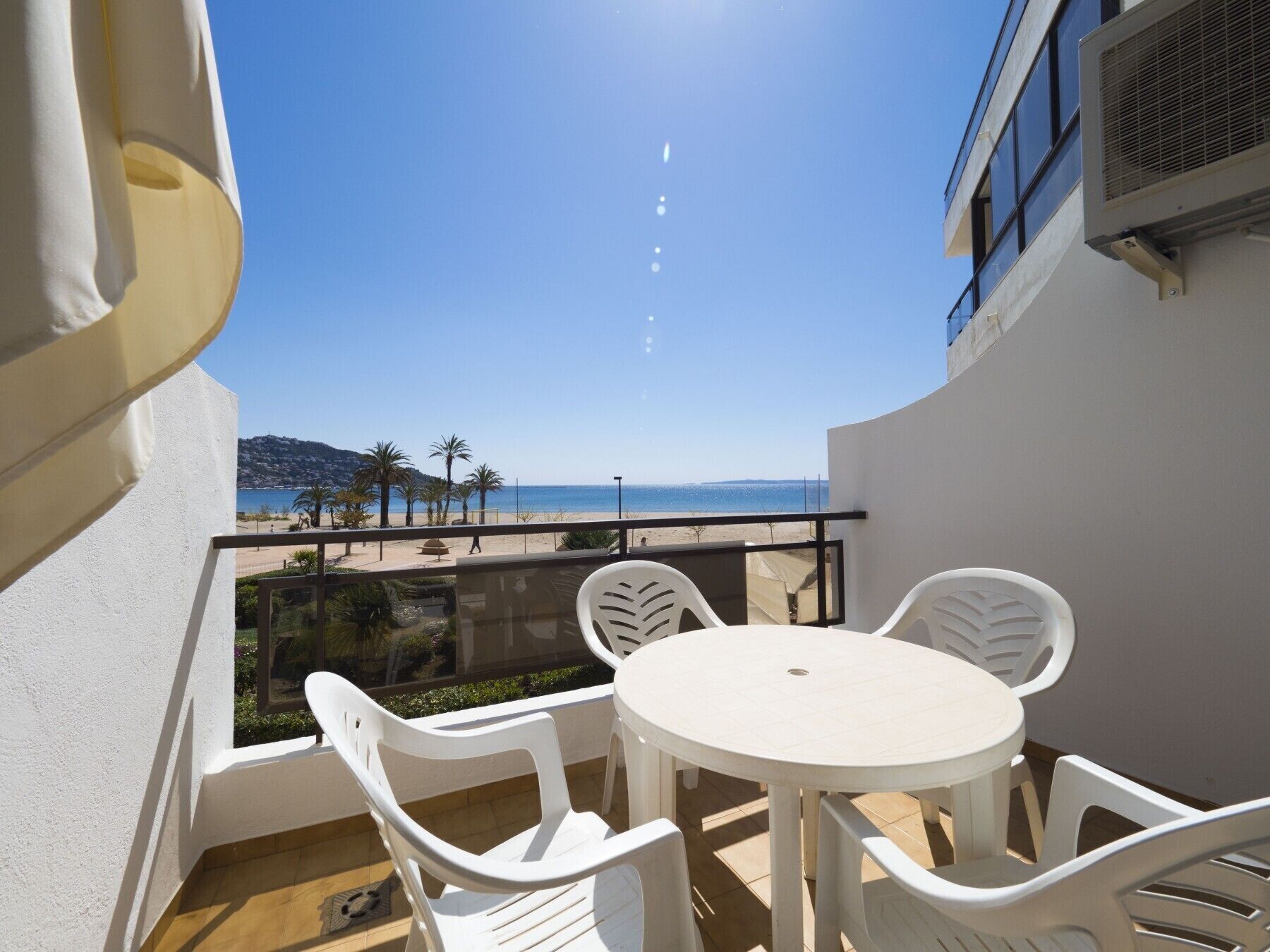 Uma varanda com mesa e cadeiras brancas, com vista para uma praia com palmeiras e céu claro.