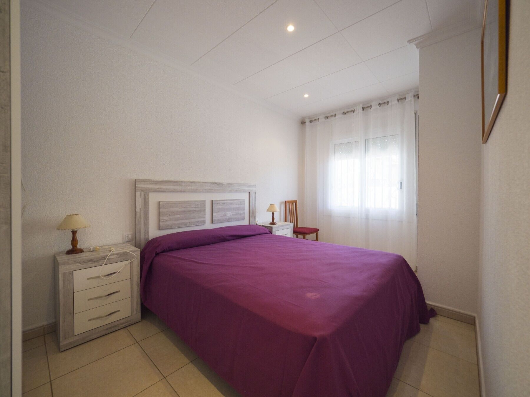 Een slaapkamer met een bed, nachtkastje, lamp, stoel en raam met gordijnen.