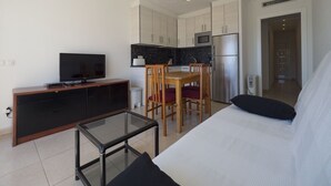 Ein kompakter Wohnbereich mit Sofa, Glastisch, Fernseher, Küchenzeile mit Schränken und Geräten sowie Esstisch mit Stühlen.