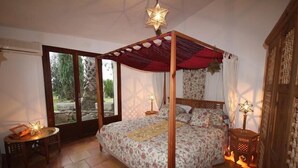 A four-poster bed with a floral bedspread, a wooden nightstand with a lamp, and a view of greenery through large sliding doors. - Large sea-view villa with 3 independent apartments (Mandelieu-la-Napoule)