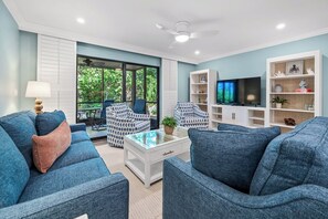 Una sala de estar con sofás azules, una mesa de centro de vidrio, un televisor y una estantería.