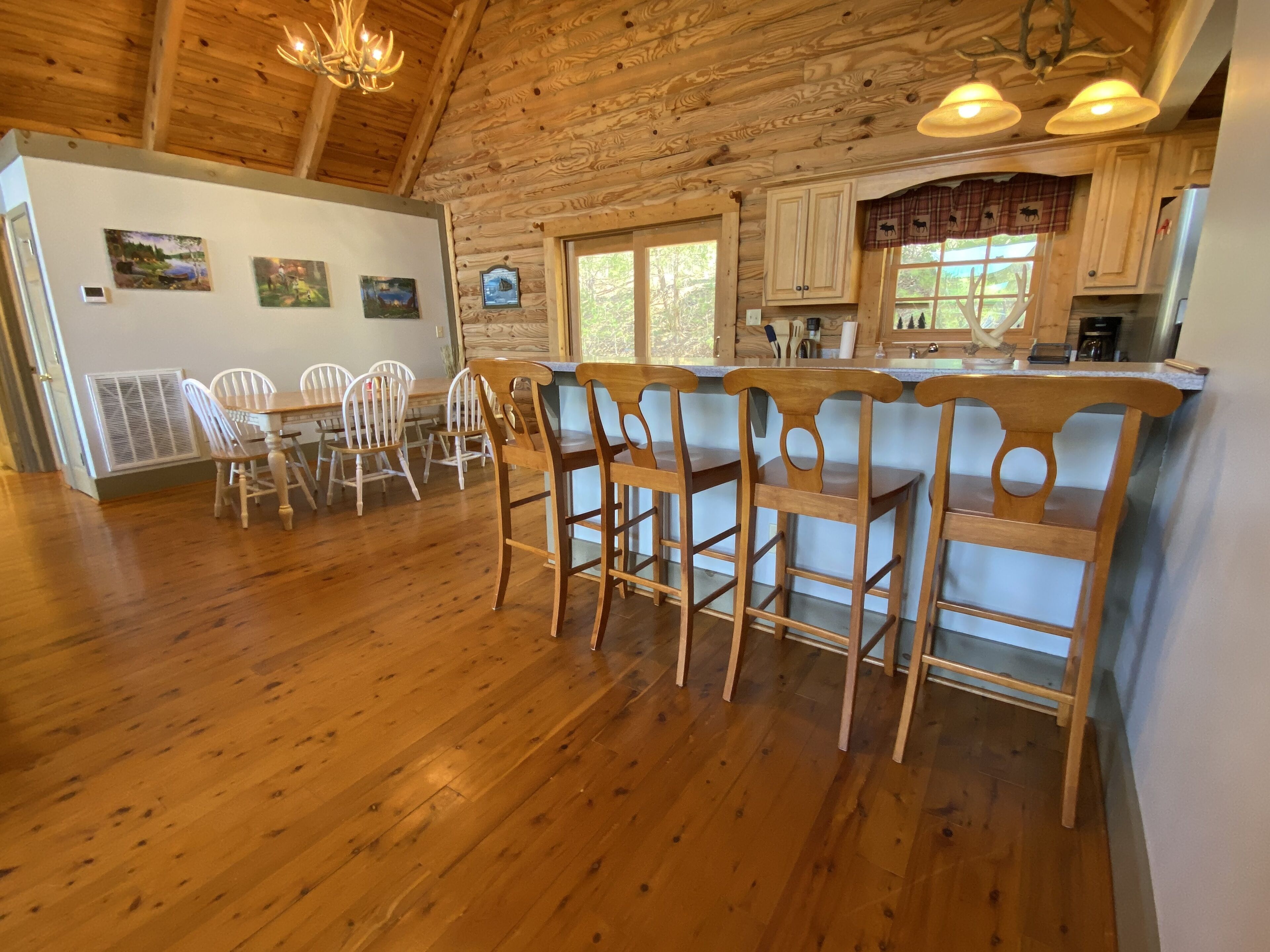 A rustic cabin interior with wooden floors, a dining area with white chairs, a kitchen with wooden cabinets, and a large window.