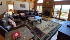 A spacious living room with a leather sofa, a coffee table, and a fireplace. There are large windows and a balcony with a view of the mountains. - Mountain Cabin,Pet-Friendly,Views,HotTub,Deck,WiFi (Ferguson)