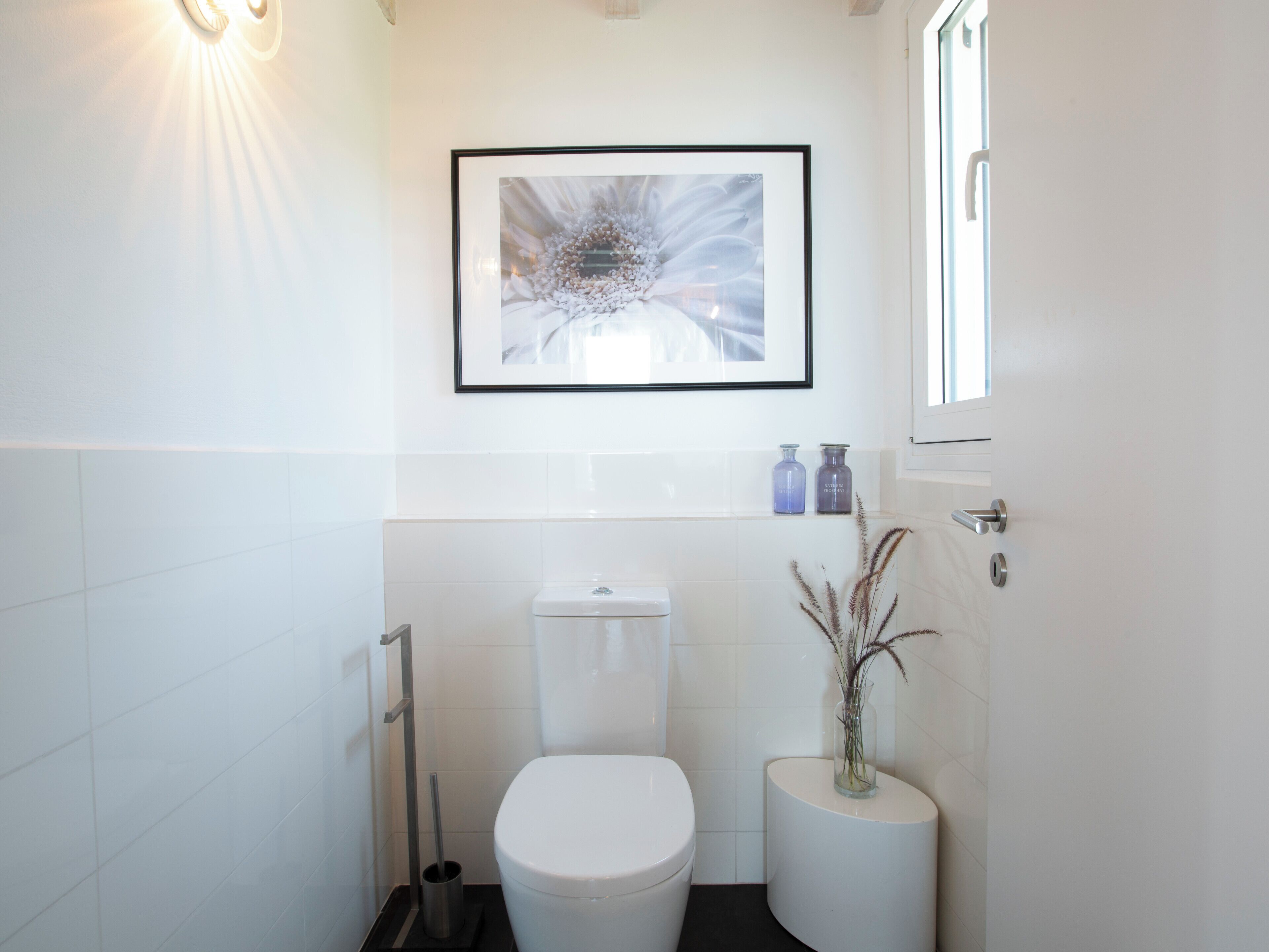 A modern bathroom with a toilet, a bidet, a framed abstract painting, and a vase with dried plants.