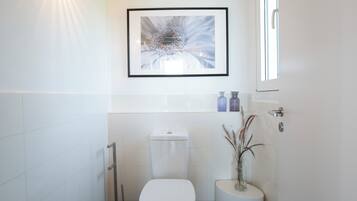 A modern bathroom with a toilet, a bidet, a framed abstract painting, and a vase with dried plants.