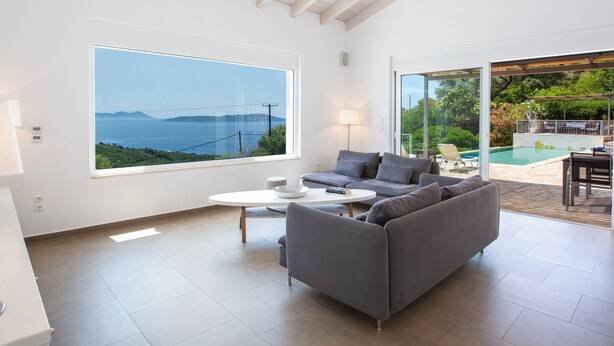 A modern living room with a large window, a sofa, a coffee table, and a view of a swimming pool and greenery.