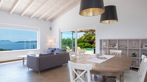 A spacious living area with a large dining table, white chairs, a grey sofa, and a view of the sea through large windows.