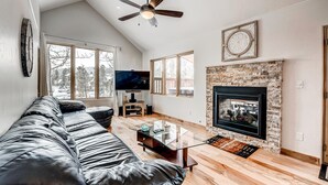 A modern living room with a black leather sofa, a flat-screen TV, a fireplace, and large windows.