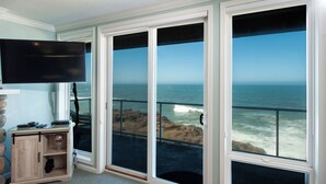 A room with a large window overlooking the ocean, a flat-screen TV mounted on the wall, and a wooden cabinet with a glass top. - Promo! Stunning Oceanfront Condo, Wifi, HDTV, Hot Tub, Indoor Pool and More! (Depoe Bay)